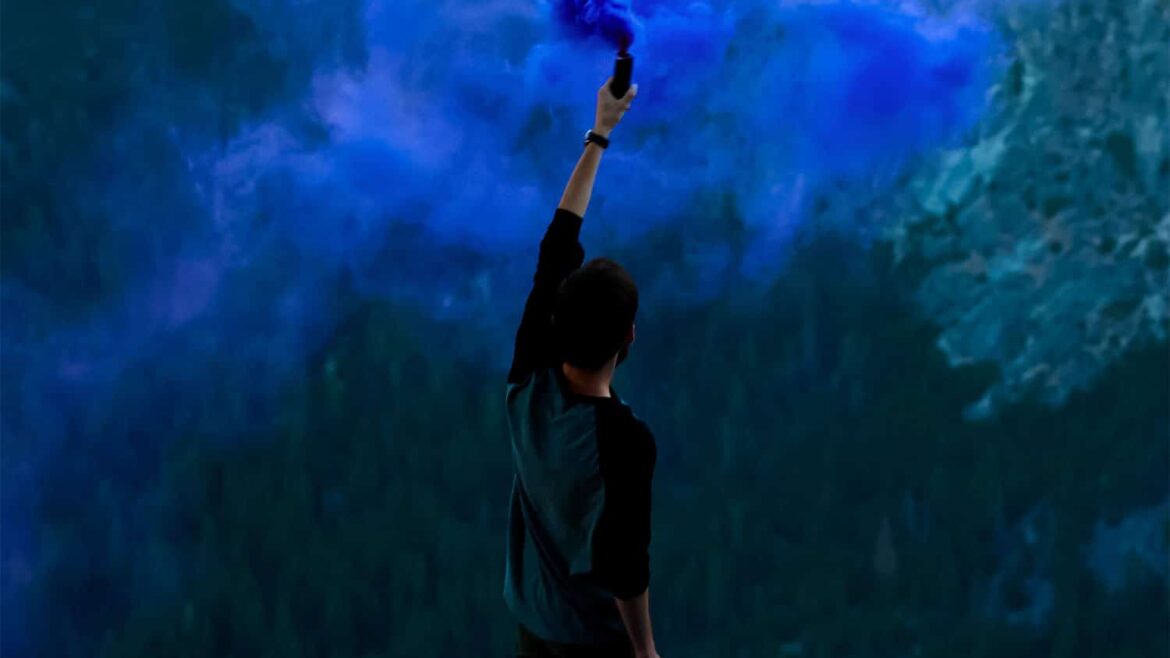 Una persona con una bengala de humo azul sobre un promontorio, al fondo se ve un paisaje montañoso