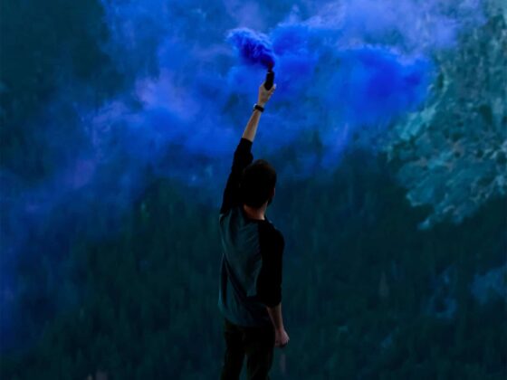 Una persona con una bengala de humo azul sobre un promontorio, al fondo se ve un paisaje montañoso