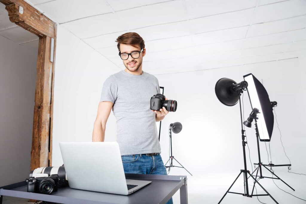 Una persona con una cámara réflex digital revisa el portátil, al fondo se ve un estudio fotográfico