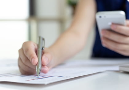 Una persona señalando con un bolígrafo en un documento con gráficos mientras examina el móvil