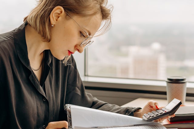 Una joven con una blusa oscura, consultando documentos con una calculadora y un bolígrafo
