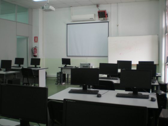 Vista interior de una aula con proyector y mesas dobles con ordenadores para cada alumno