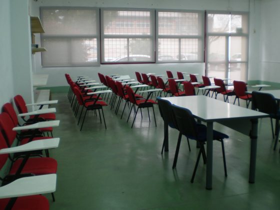 Vista interior de un aula dónde vemos varias mesas dobles y sillas individuales con pupitre