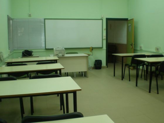 Vista interior de un aula dónde vemos varias mesas dobles, la mesa del docente y la pizarra blanca