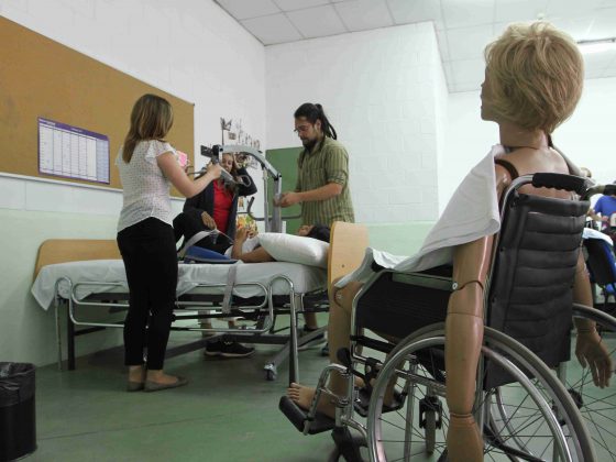 Varias personas aprenden a movilizar a personas dependientes, delante un maniquí en silla de ruedas